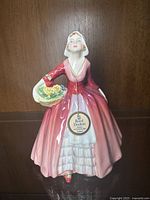 Front view of Royal Doulton figurine Janet wearing a gradient pink-red dress holding a basket of yellow roses with original tag hanging