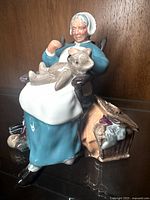Front and side view of Royal Doulton Nanny figurine showing the elderly woman in a blue dress and white bonnet seated with a teddy bear on her lap and picnic basket beside the rocking chair.