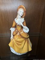 Front and side view of Royal Doulton 'Sandra' porcelain figurine in yellow dress and bonnet, seated pose