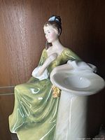 Front-side angle of Royal Doulton 'Secret Thoughts' porcelain figurine showing seated woman in green dress next to a white column holding a hat with flowers.