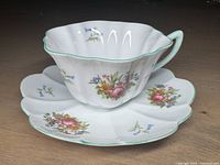 White scalloped Shelley cup and saucer with green trim and floral patterns, placed on a wooden surface.