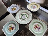 Five vintage ceramic plates arranged on glass table showing colorful floral and fruit patterns with scalloped edges and textured rims.