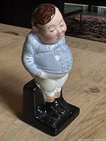 Front side view of the vintage Royal Doulton Fat Boy figurine showing the boy's face, blue jacket with button details, white pants, peach socks, and black boots on black rectangular base.