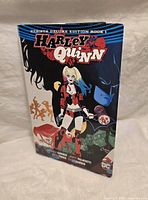 Front cover of Harley Quinn Rebirth Deluxe Edition Book 1, showing Harley Quinn with supporting characters Batman and others. The book is hardcover and displays vibrant comic art.