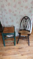 Photo of a green wooden stool with a woven wicker seat next to a dark wooden chair with a woven wicker seat and decorative spindled backrest, placed on hardwood floor with floral wallpaper background.