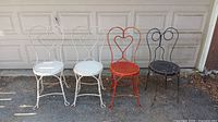 Four metal cafe chairs arranged in a row, showing two white heart swirl back chairs, one red heart swirl back chair, and one black circular swirl back chair on a concrete surface in front of a garage door.