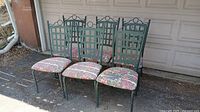 Photo showing six green metal outdoor chairs with lattice-style backs and floral seat cushions arranged in a row outdoors.