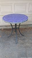 Full view of the round outdoor table with blue tile top and black metal legs on an outdoor surface.