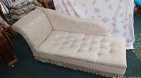 Side angled view of cream chaise longue showing curved side arm, tufted seat and fringed base