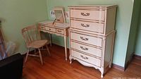 Photo showing the full set of a cream and gold vanity, dusty pink-beige chair, and matching five-drawer dresser in a room with hardwood floors.