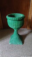 Front angled view of green wicker planter showing rounded bowl top, twisted pedestal base, and square bottom against brown wood background.