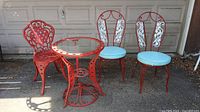 Outdoor set featuring two red metal chairs with white ornate backrests and blue cushions, one additional matching chair, glass top table, all with some rusting.
