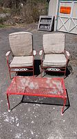Photo of two metal chairs with beige cushions and a red metal mesh coffee table outdoors showing overall condition with rusting.