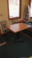 Photo of wood-topped table with three-legged black iron stand and two metal chairs with mosaic backs beside a window.
