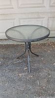 Full view of round outdoor table with metal frame and glass top placed outdoors on gravel surface.