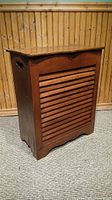 Front view of wooden laundry hamper showing slatted front and warm brown finish against wood-paneled background.