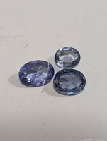 Three oval cut genuine tanzanite gemstones displayed on a plain surface showing clarity, color, and cut.
