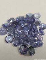 Close-up photo showing 27 loose oval cut tanzanite gemstones in varying shades of blue-violet on a white surface.