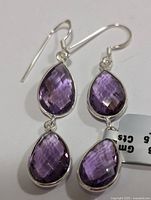 Close-up photo of a pair of sterling silver drop earrings, each featuring two faceted pear-shaped purple amethyst stones set in silver frames with hooks.