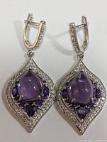 Front view of a pair of silver earrings with a central purple pear-shaped amethyst surrounded by smaller amethyst and cubic zirconia stones