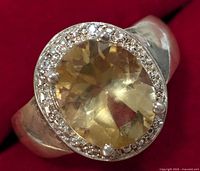Close-up photo of silver ring with an oval citrine surrounded by small clear stones on a red background.