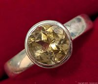 Close-up image of a silver ring with a round faceted citrine gemstone set in a bezel setting on a red background, showing the faceted gemstone and silver band.