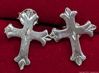 Close-up of two silver cross-shaped earrings on a red background, showing front side with decorative ends.