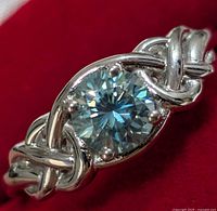 Close-up front view of silver ring with round moissanite stone and woven band design on red background.