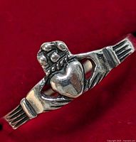Close-up photo of a silver Claddagh ring on a red background showing a heart held by two hands with a crown above.
