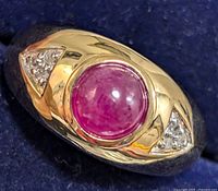 Close-up image of a silver ring with a round cabochon ruby center stone and clear pavé side stones on a blue background.