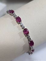Close-up photo of silver bracelet featuring oval red rubies and round diamond accents on white display cushion.