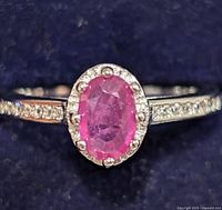 Close-up photo of silver ring showing oval pink ruby center stone with surrounding diamond accents, on a detailed band with small diamonds.