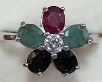 Front close-up of floral cluster ring showing oval ruby, emeralds, blue sapphires, and central round white stone
