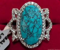 Close-up top view of silver ring with large oval turquoise stone and surrounding small clear stones, showing intricate silver detailing.