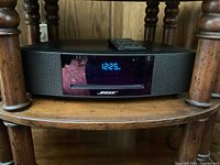 Front view of Wave Music System on wooden shelf showing digital clock display reading 12:25 PM with remote on top