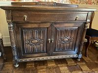 Front view of wooden cabinet showing carved front doors, side carved columns, metal handles, and top drawer.