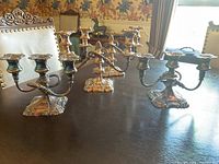 Three silver tone candelabras on a dark wood table showing overall shape, detailing and size relative to chairs