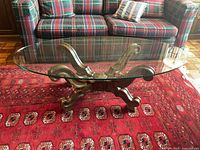 Glass top coffee table with ornate wooden base on red patterned rug and plaid sofa background.