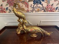 Pair of gold-colored wood bird sculptures, one upright on curved base and one elongated on pedestal base, on a wooden surface with floral wallpaper background.
