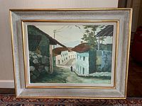 Front view of a framed landscape painting depicting a quiet village street scene with buildings and a figure.