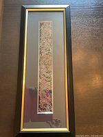 Full view of framed embroidery with black and gold frame, showing narrow vertical panel of floral embroidery on deep background.