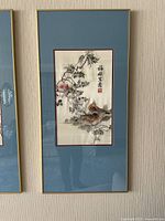 Framed artwork of embroidered fabric with birds and floral branches and Chinese calligraphy, mounted with blue mat and gold frame.