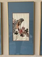 Framed embroidered fabric art piece depicting a detailed bird with colorful feathers and floral branch on cream fabric background inside a blue mat and gold frame.