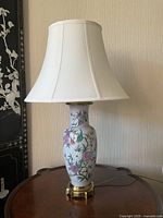 Full view of the porcelain lamp sitting on wooden furniture, showing the floral design, white lampshade, and brass base.