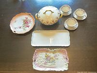 Overview of all items in the lot on a wood surface showing serving bowl, tea set (3 cups and saucers), rectangular white tray, and two decorative porcelain trays with pink floral designs