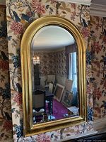 Front view of a large wall mirror with gold leaf arched frame hung on floral wallpaper.