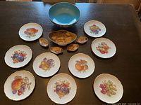 Top-down photo showing a collection of fruit-themed porcelain plates and bowls spread on a wooden surface. Plates have gold scalloped edges with colorful fruit illustrations; three small oval bowls with yellow interior and fruit patterns; one larger blue interior bowl.