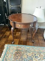 Front view of wooden side table showing scalloped top edge, drawer with brass handles, and cabriole legs.