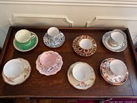 Eight teacup and saucer pairs arranged on wooden tray, overhead view