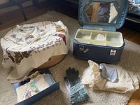 Blue vintage suitcase opened showing broken mirror inside lid and compartments, multiple folded scarves on the floor, a small pair of black leather gloves in packaging, and a vintage pink hat.
